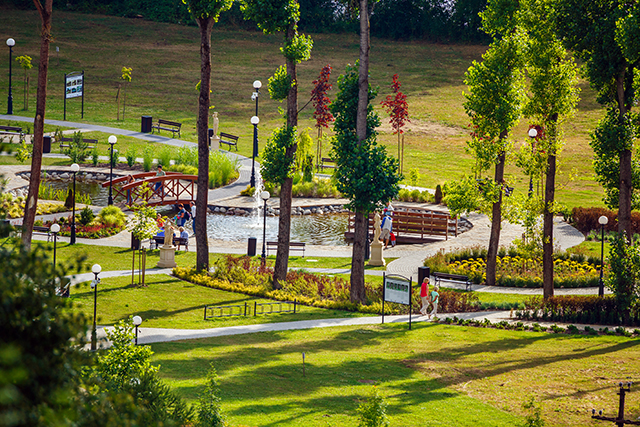 Zdjęcie przedstawia widok ogólny parku pełnego ścieżek, oczek wodnych i kwiatów, a także spacerujących ludzi.