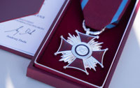 Powiększ zdjęcie: Bliskie zdjęcie odznaki "Srebrny Krzyż Zasługi" - krzyż na promienistym tle w ciemnoczerwonym pudełku. 